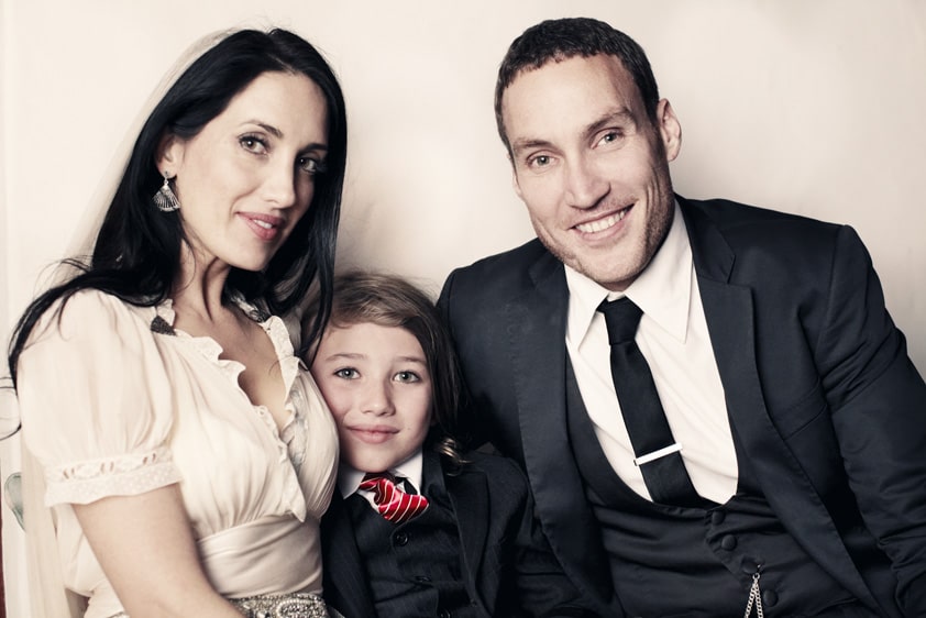 Callan Mulvey in a black formal suit with his wife in a nude color dress and son in a black suit.